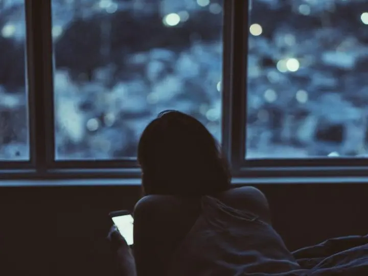 Foto: eine Person liegt nachts am Fenster und schaut aufs Handy, draußen die Lichter der Stadt